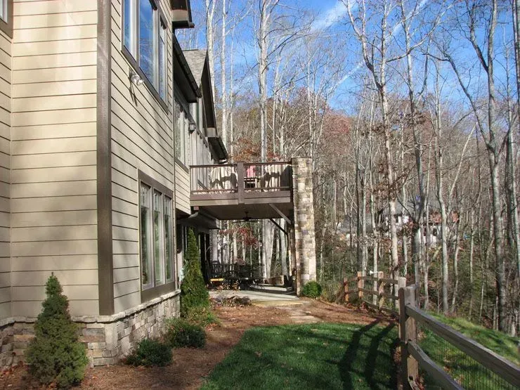 Beige house with a deck overlooking a wooded area. Brown fence and fallen leaves in the grass.