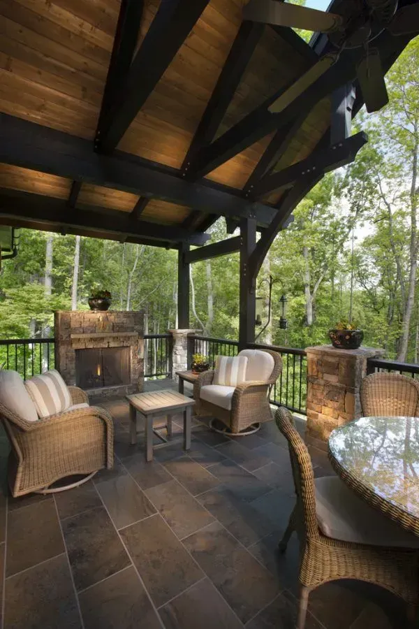 Patio with wicker furniture, fireplace, and overhead wooden beams overlooking a lush green forest.