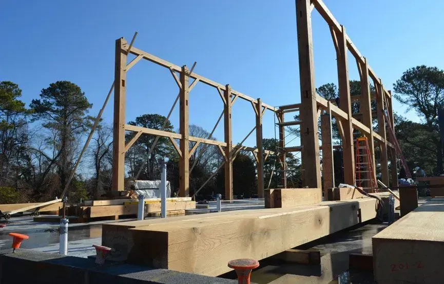 Wooden frame of a building under construction on a concrete foundation, outdoors with trees.