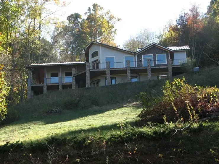 House on a hillside with a deck and stone accents, surrounded by trees and grass, on a sunny day.