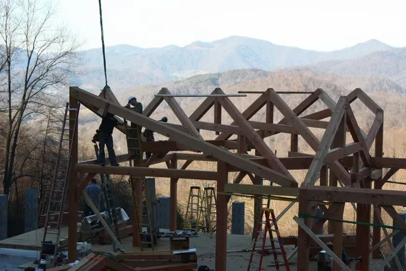 Construction workers assembling a wooden timber frame building on a hillside, with mountains in the background.