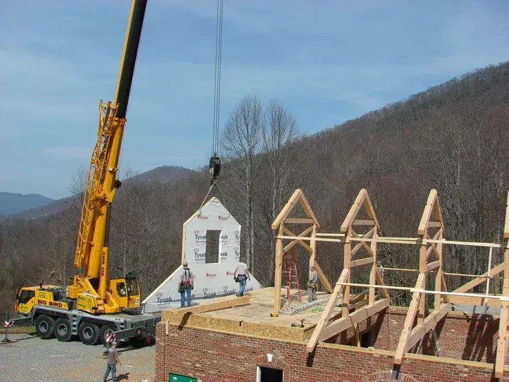 A crane lifting a prefabricated wall section onto a building under construction, with workers and a mountain backdrop.