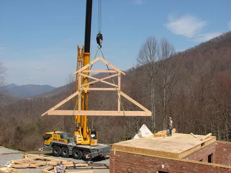 A crane lifts a wooden roof truss to be placed on a brick building under construction with a mountain backdrop.