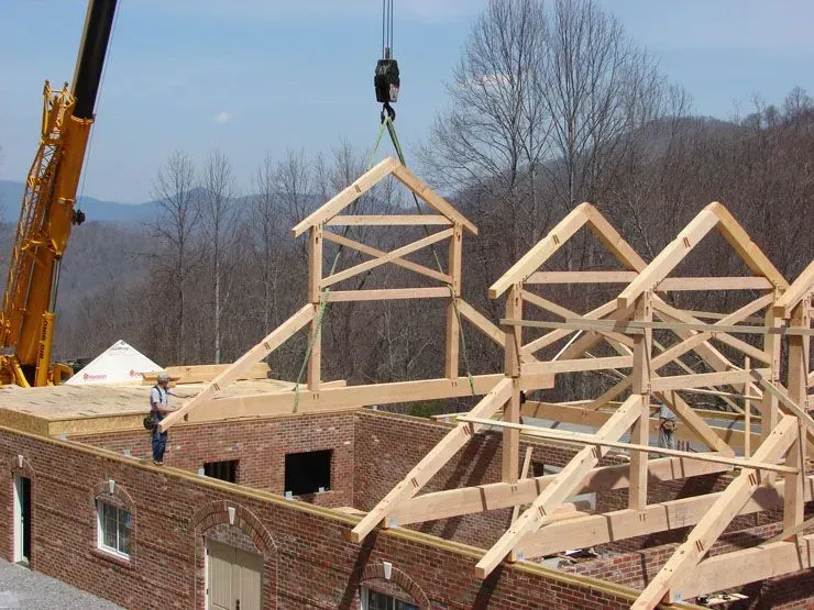 Crane lifting wooden truss onto a brick building under construction with a mountainous background.