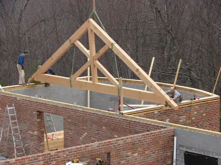 Construction workers positioning a wooden roof truss on a brick building's curved wall.