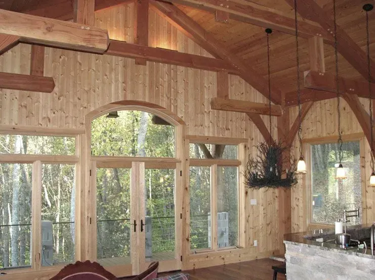 Interior of a rustic room with wooden walls, large windows, exposed beams, and a view of trees.