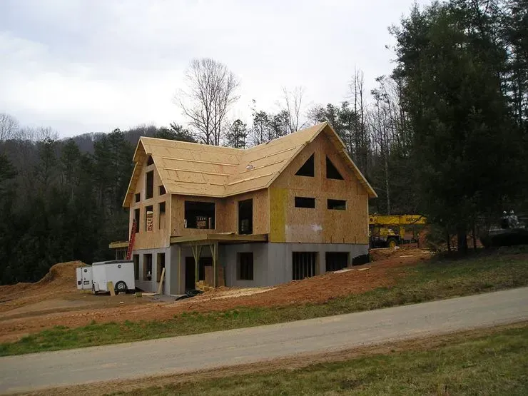 House under construction; wood frame with unfinished exterior, nestled in a wooded hillside, near a road.