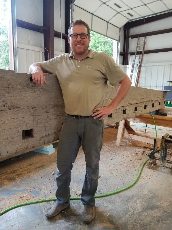 Man in a tan shirt and gray pants smiles, leaning on a large wooden beam in a workshop.
