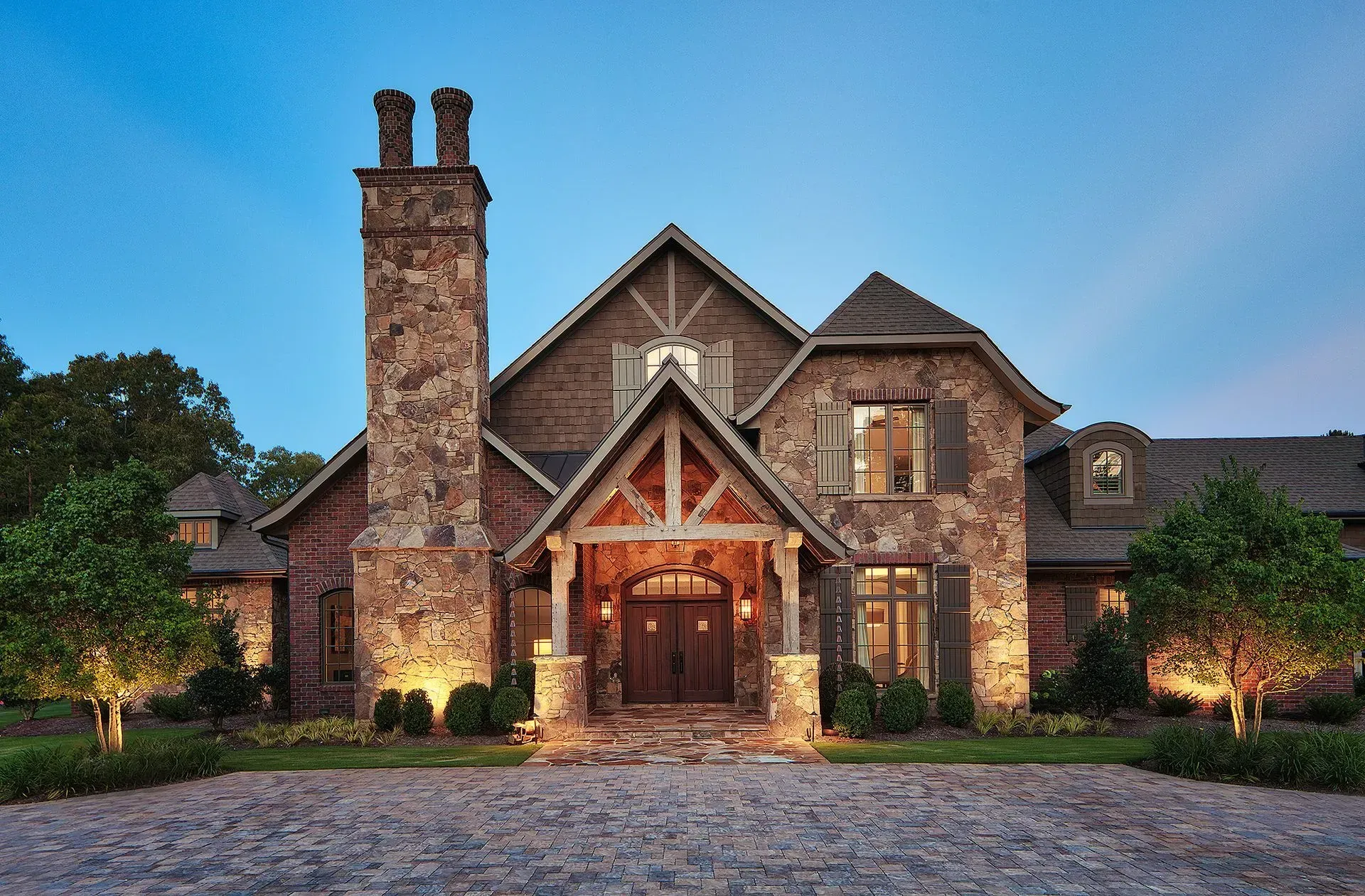 Stone house with a large chimney, wooden accents, and a cobblestone driveway at dusk.