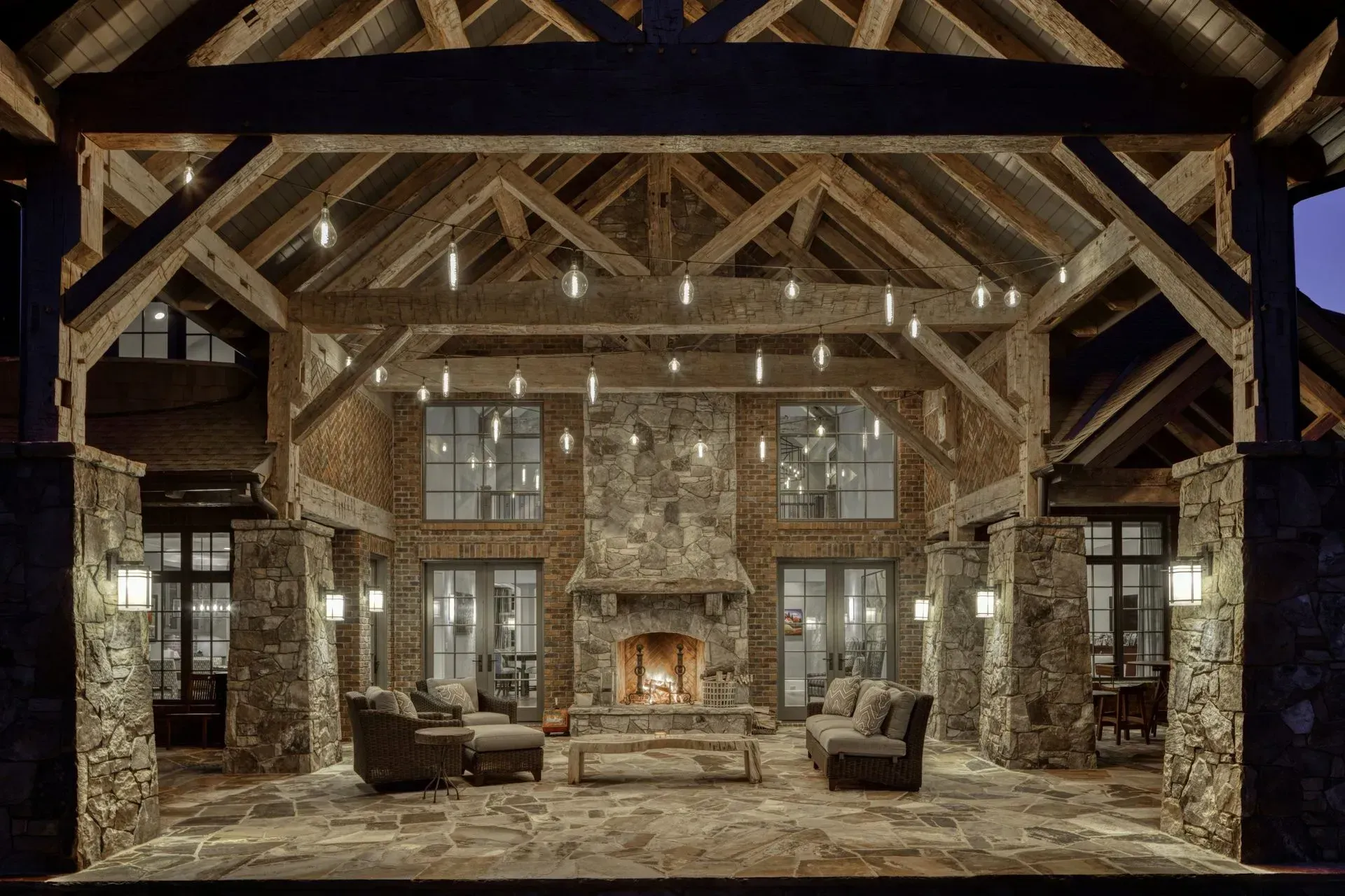 Rustic outdoor living room with fireplace, stone pillars, wooden beams, and seating.