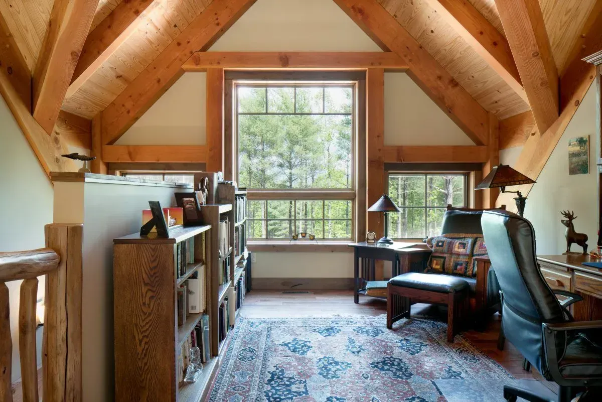 Cozy loft office with wooden beams, large window overlooking trees, bookshelves, desk, and armchair.