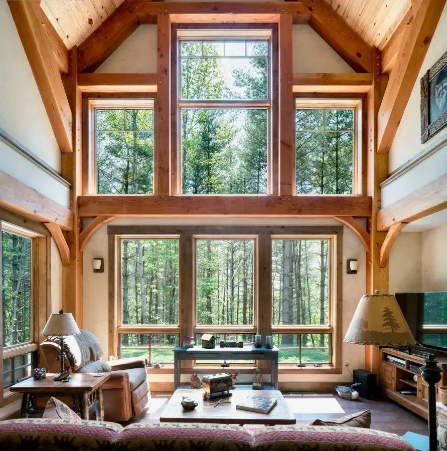 Living room with large windows, wooden beams, forest view. Furnishings: sofa, chairs, lamps.