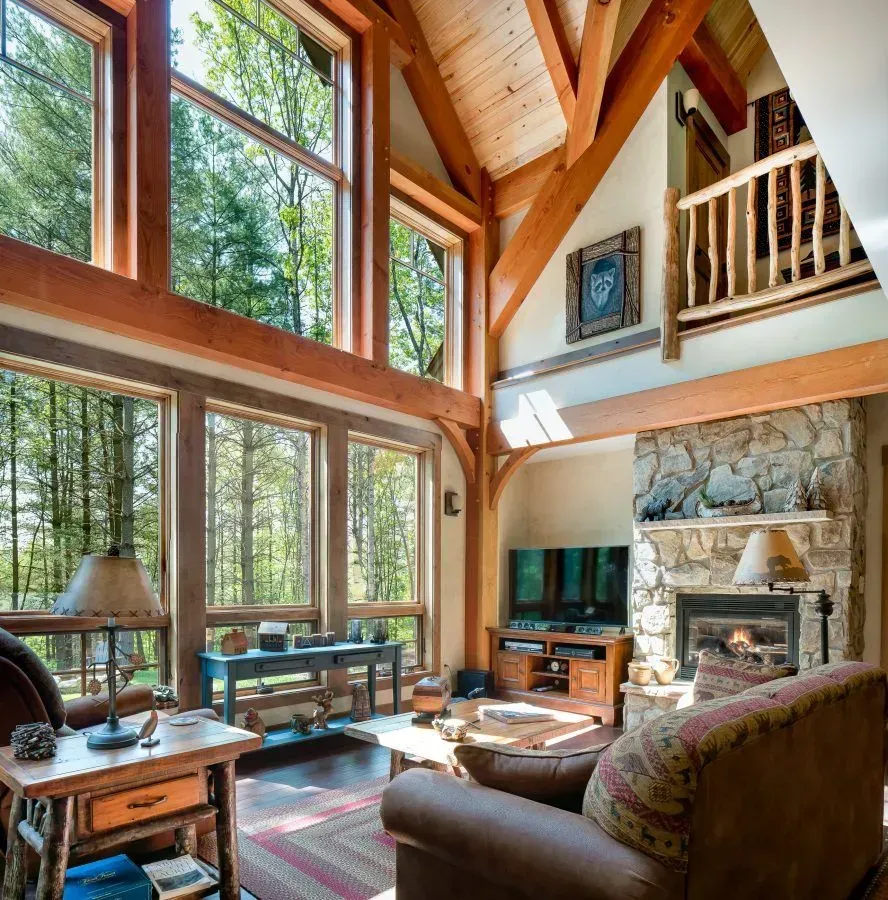 Living room with large windows overlooking a forest, stone fireplace, wooden beams, and cozy furniture.