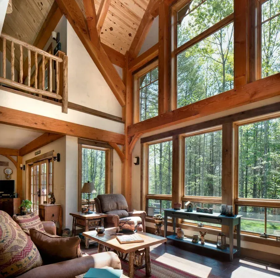 Rustic living room with exposed wood beams, large windows overlooking a forest, and a loft.
