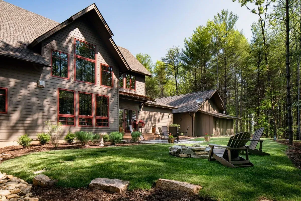 Brown two-story house with large windows, surrounded by green lawn, trees, and Adirondack chairs.