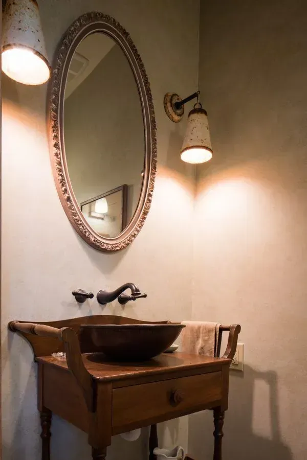 Bathroom with a wooden vanity, bowl sink, oval mirror, and sconces; neutral tones.