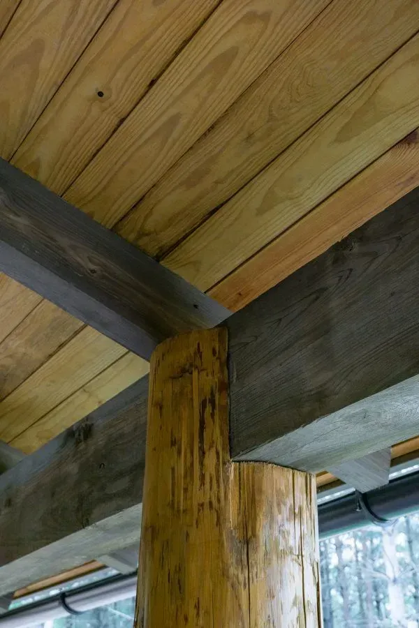 Wooden beams support a wood-plank ceiling. A dark gray beam meets a natural wood column.