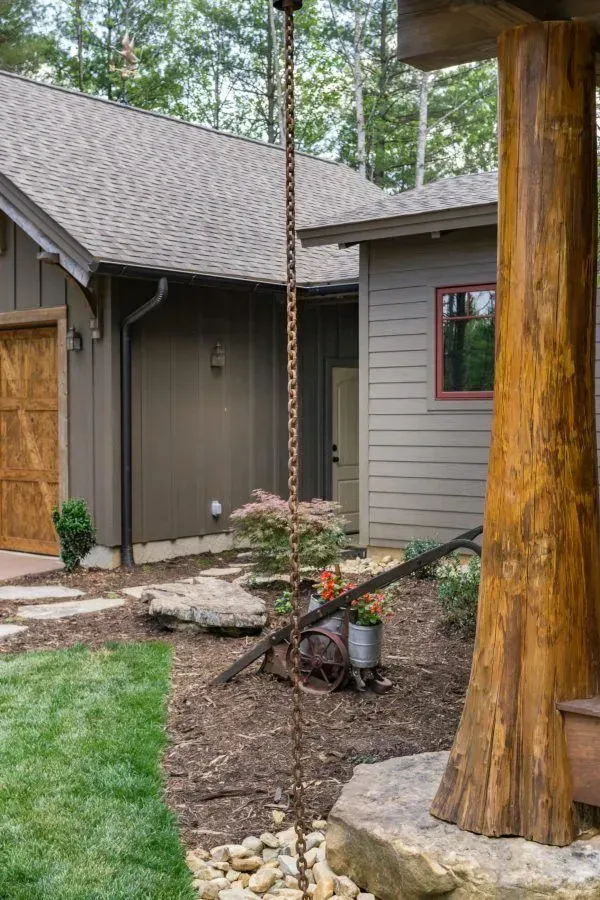 Rain chain draining into a bed of rocks near a brown house with a wooden support beam.