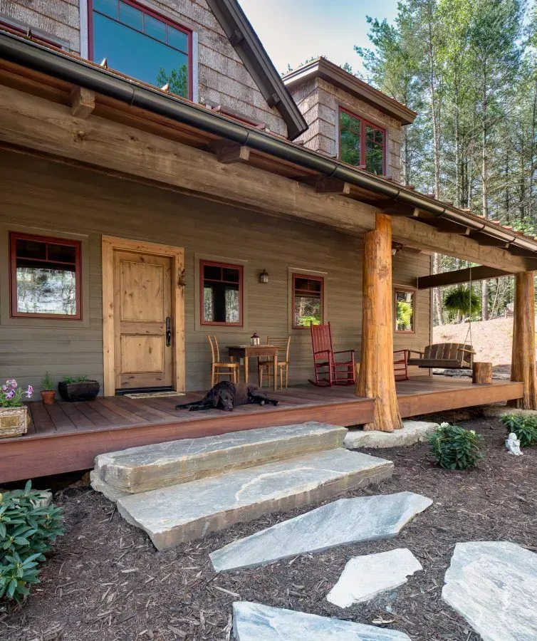 Rustic cabin with a wraparound porch and stone steps. A dog rests on the porch.