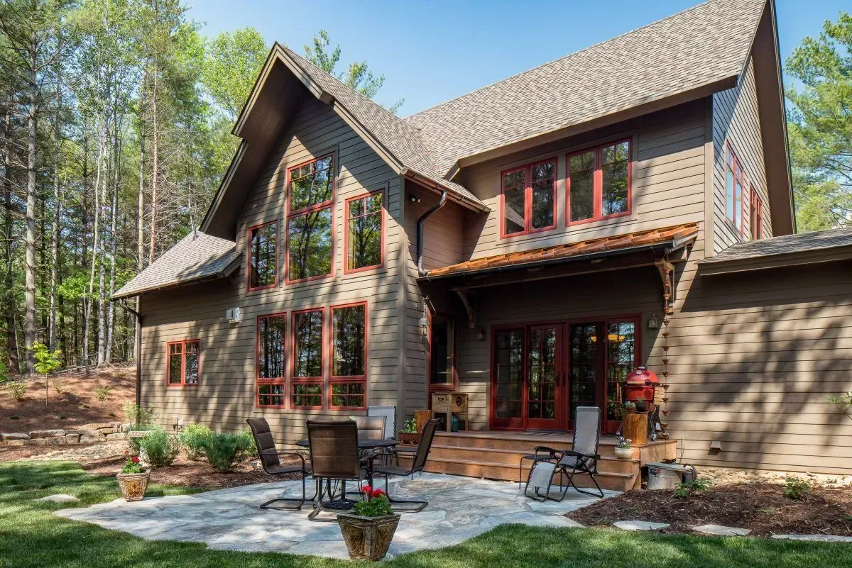 Brown house with large windows, red trim, and a stone patio in a wooded area.