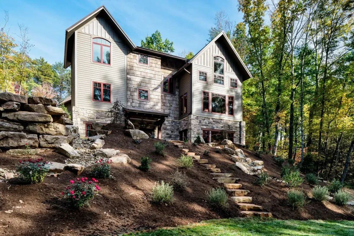 Stone house on a hillside with stairs leading up, surrounded by trees and landscaping, under a blue sky.