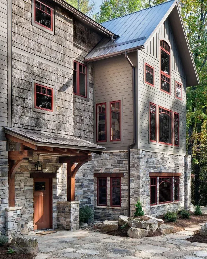 Stone and shingle house with red-framed windows and wooden porch, in a natural setting.