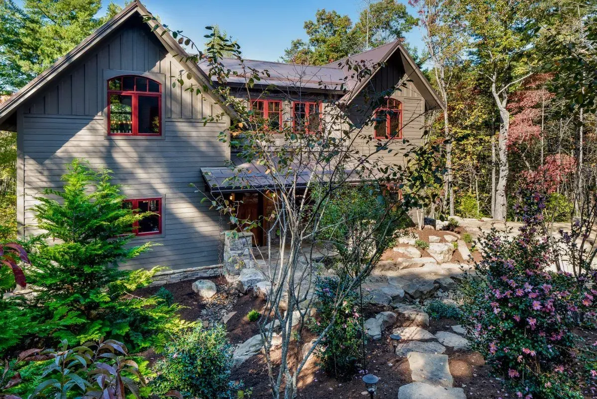 Gray house with red-trimmed windows nestled in a wooded landscape, stone path leads to the entrance.