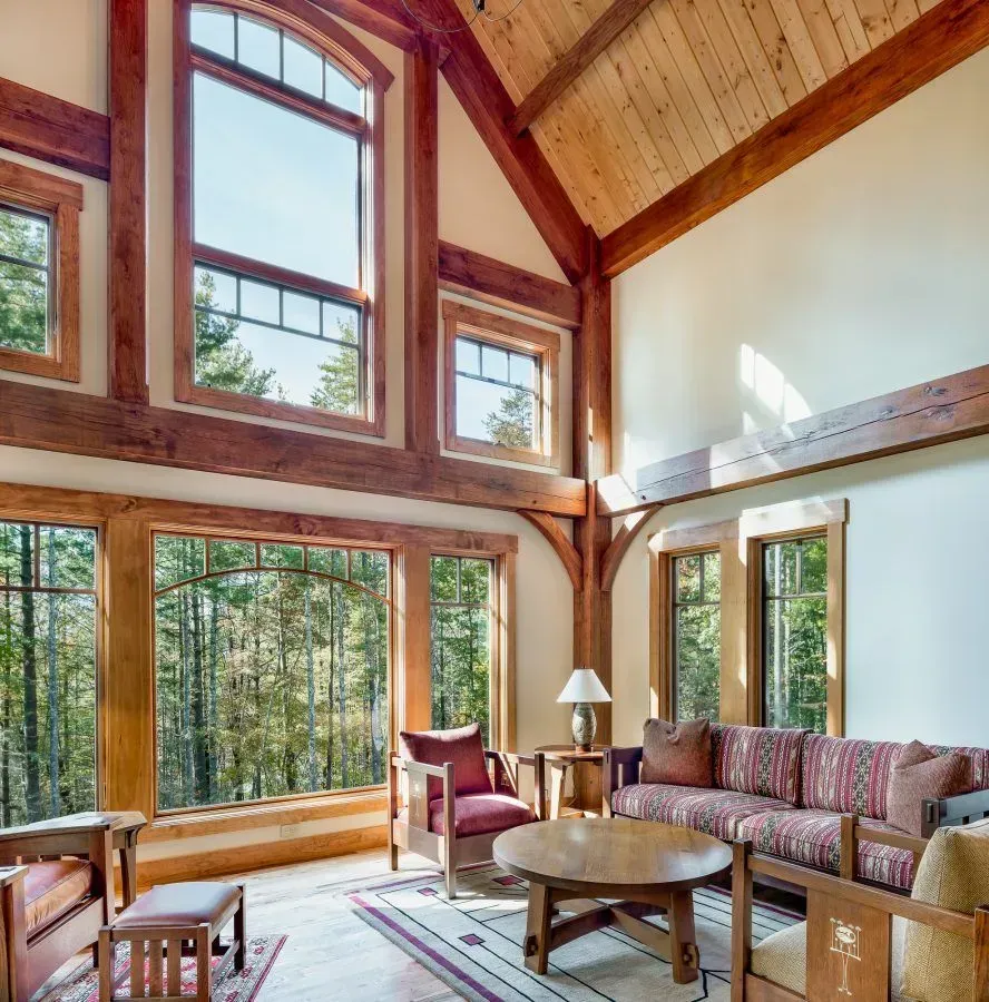 A spacious living room with large windows overlooking a forest, featuring wood beams, a sofa, and armchairs.