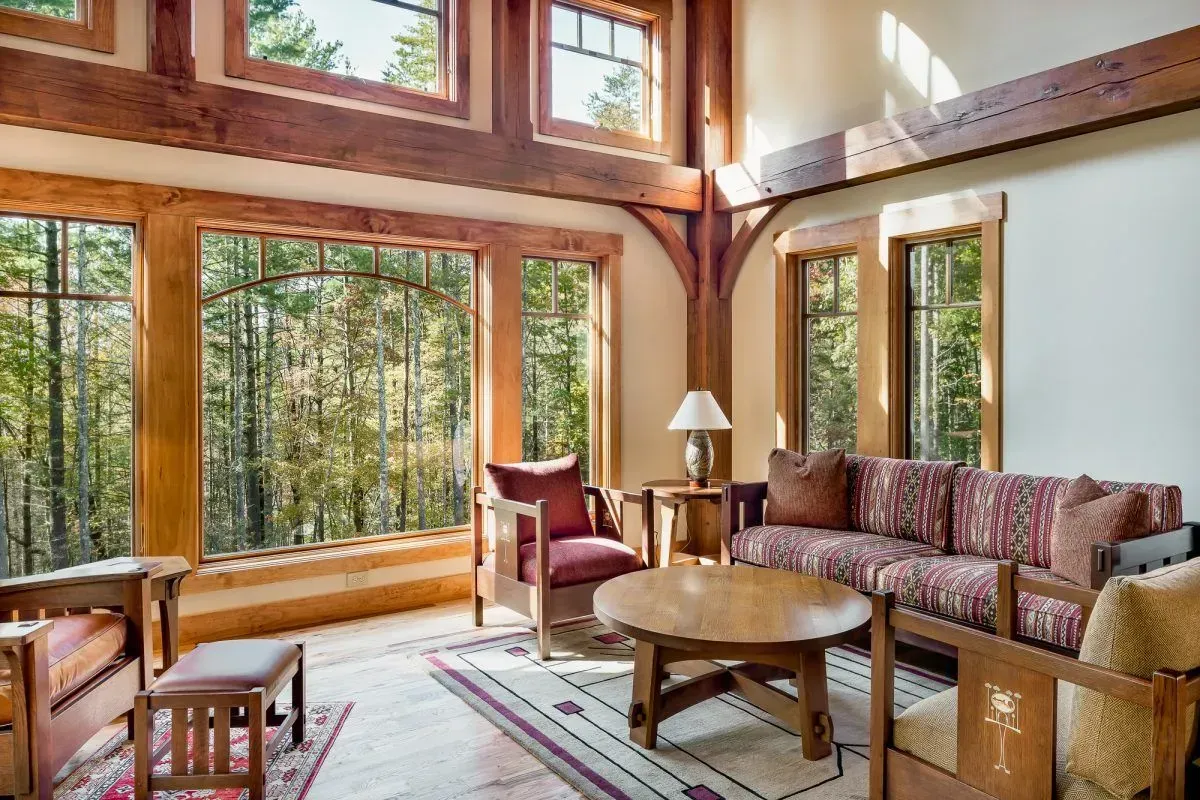 Living room with large windows overlooking a forest; wooden beams and furniture, red accents.