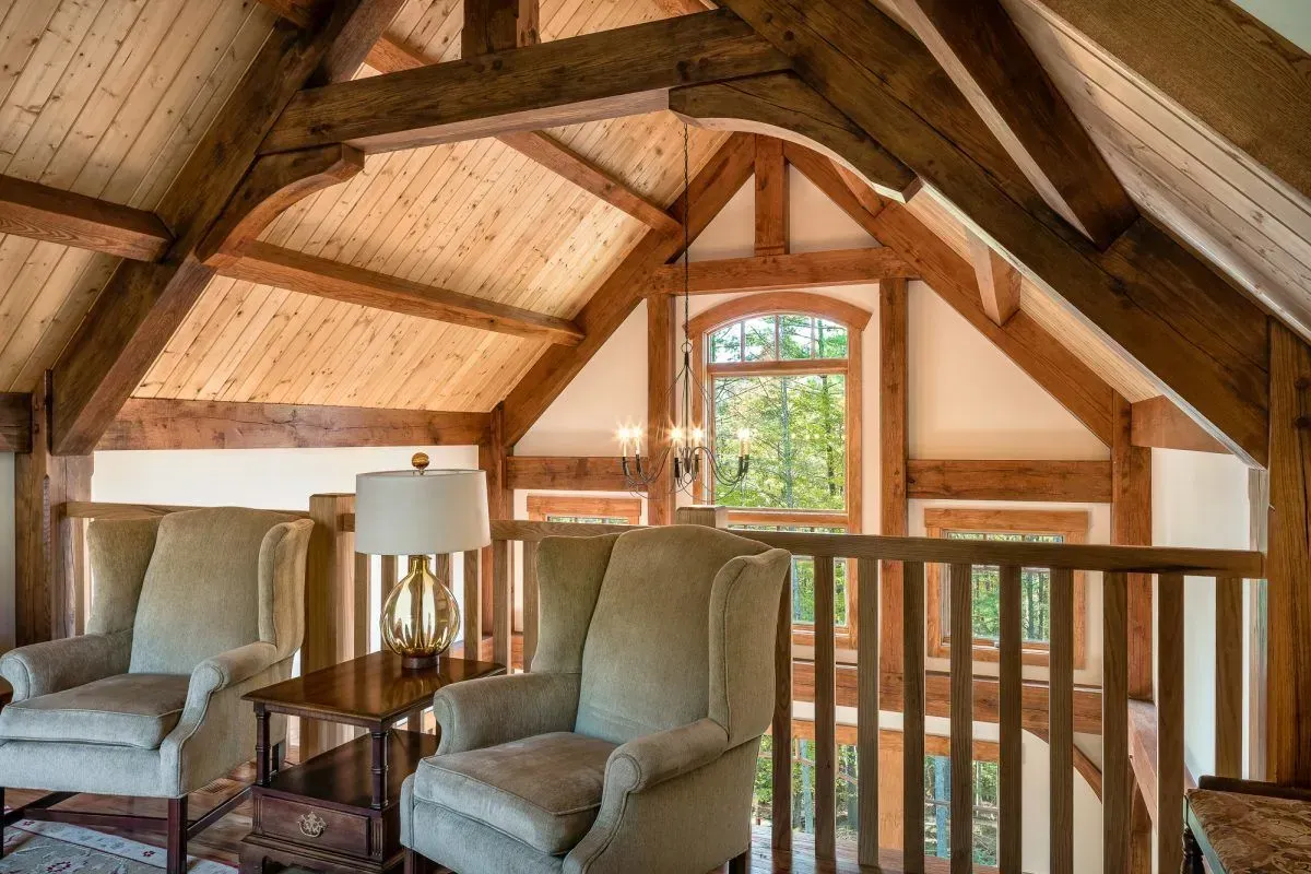 Two beige armchairs in a loft, with exposed wooden beams, a window, and a railing.