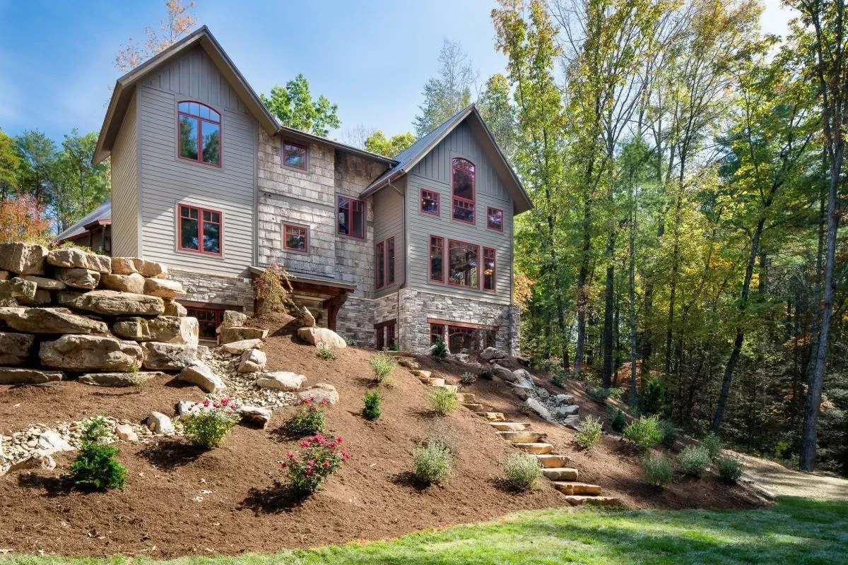 Two-story house with stone and wood exterior on a hillside, surrounded by trees and landscaping.