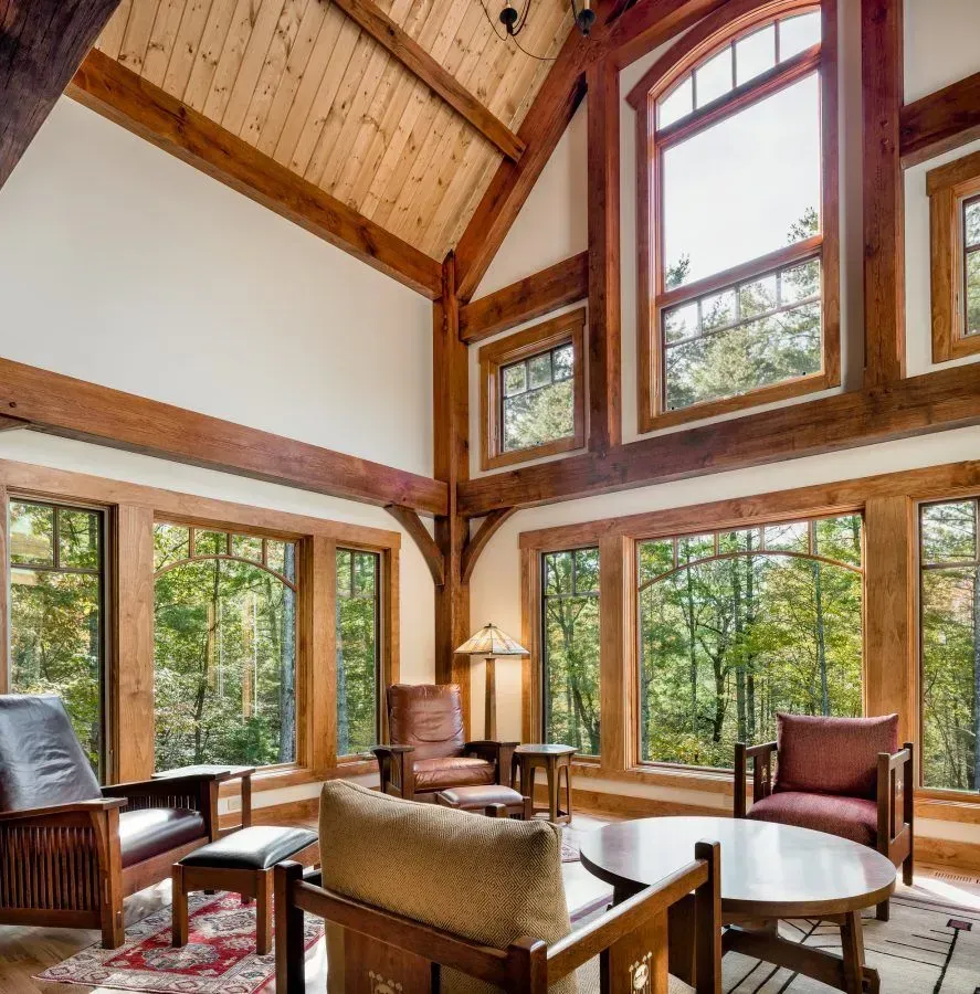 Living room with wood beams, large windows, and leather furniture overlooking a forest.