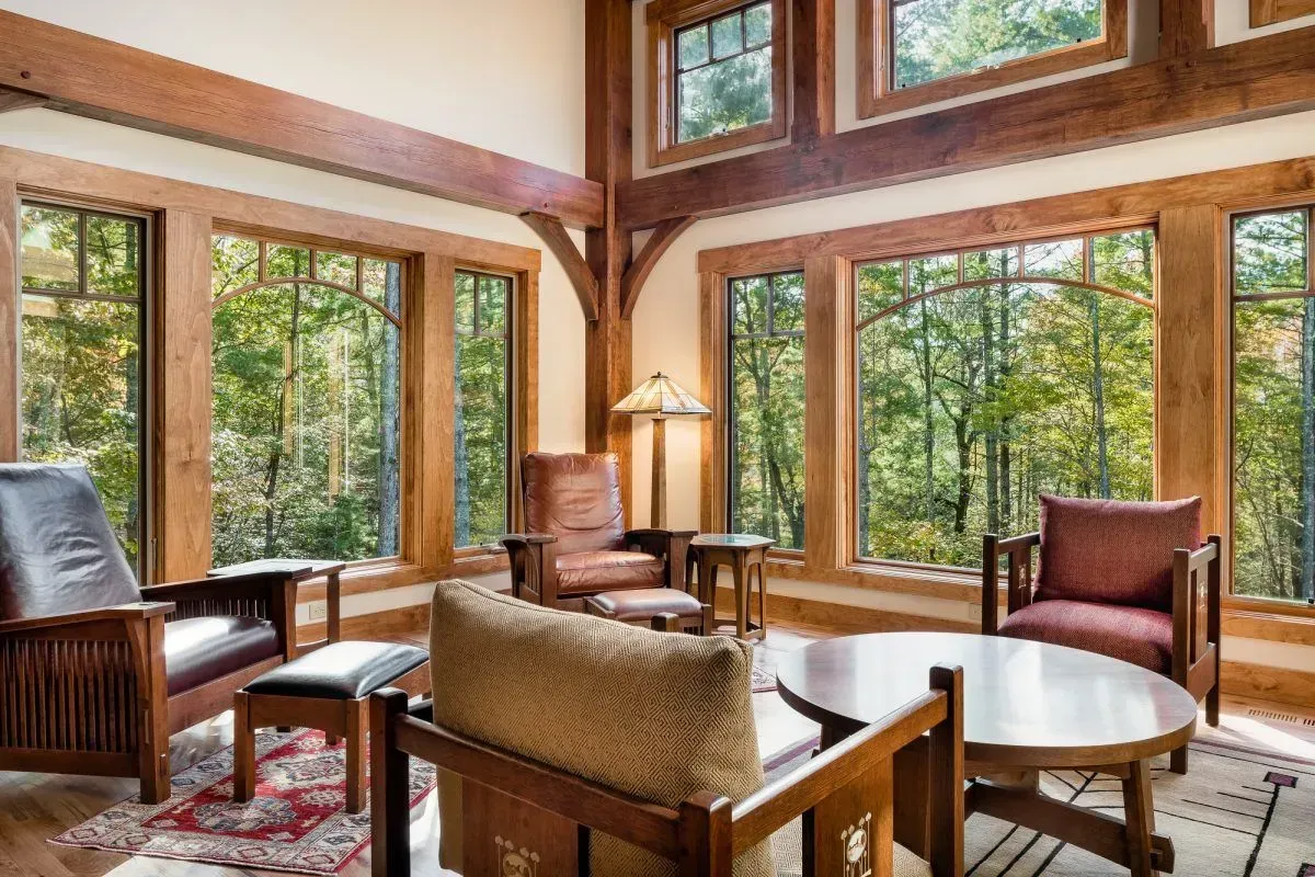 Living room with large windows overlooking a forest; wooden beams, chairs, and tables.
