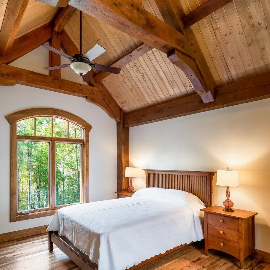 Bedroom with wooden ceiling, bed, nightstands, arched window overlooking trees.