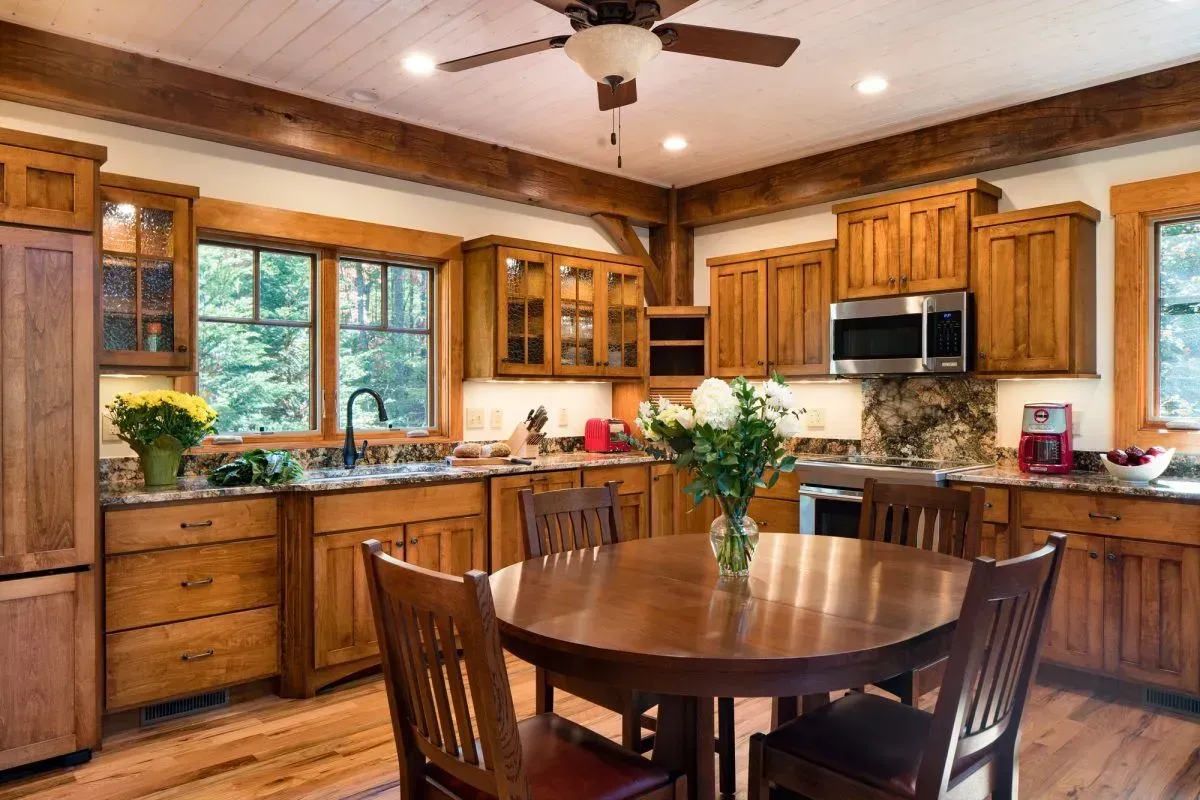 A rustic kitchen with wood cabinets, countertops, and a round table. A ceiling fan and windows are present.