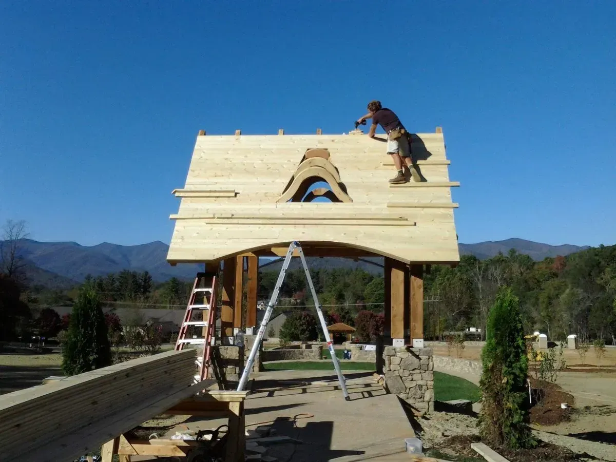 Man building roof on a wooden structure, set against a mountain backdrop under a blue sky.