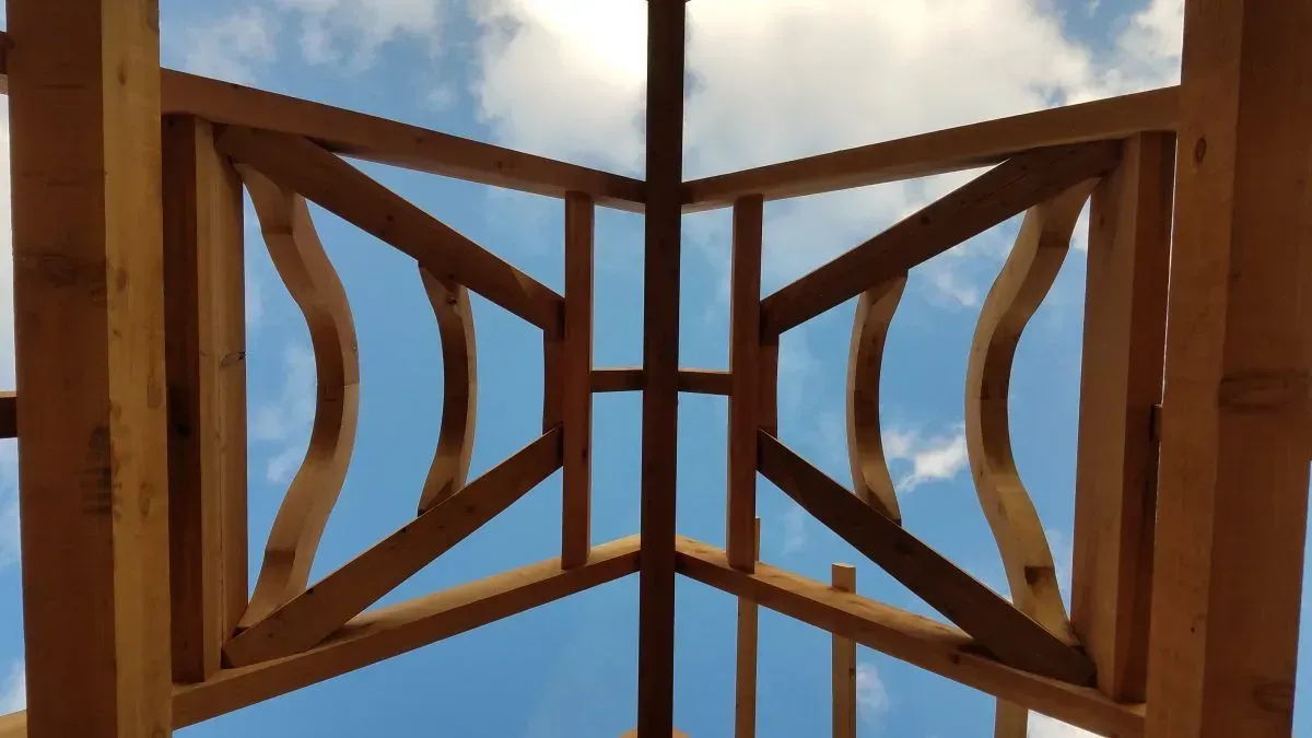 Wooden roof framework against a blue sky, featuring curved beams and rectangular supports.