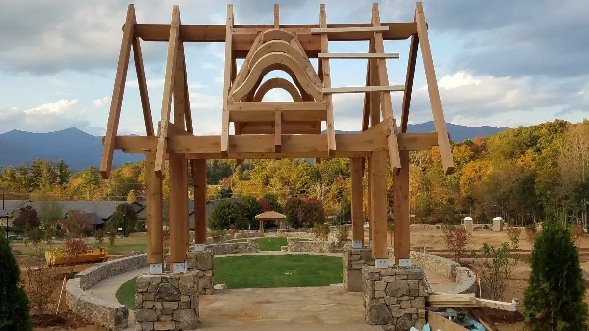 Wooden structure in a garden, under construction.  Brown beams, stone base, with mountains and autumn foliage in the background.