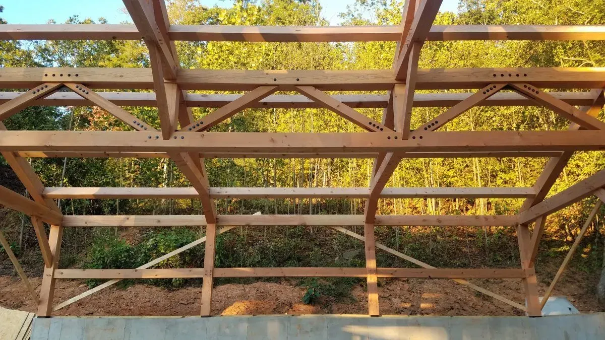 Wooden frame structure with trusses, against a backdrop of trees and sky.