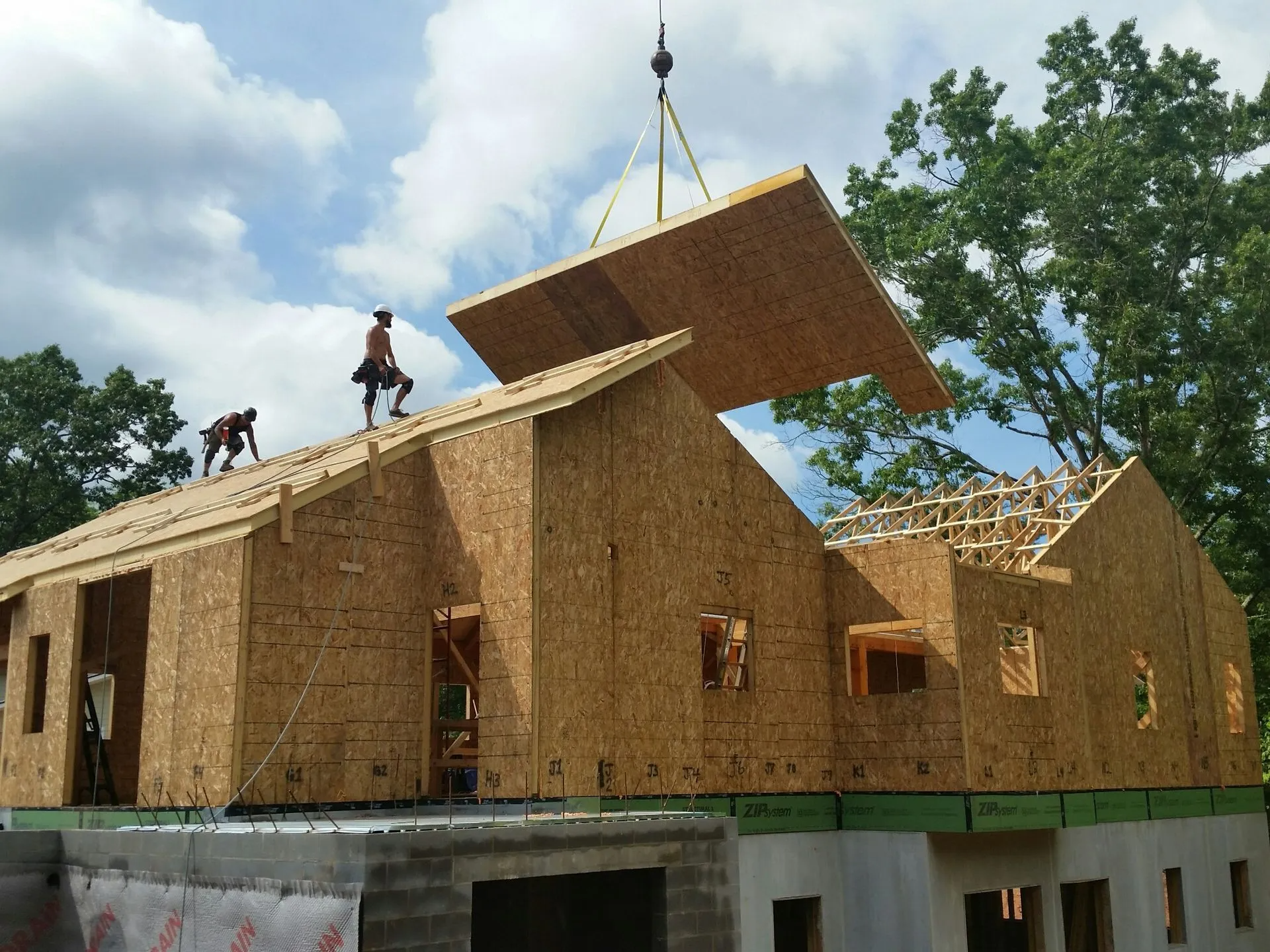 Construction workers installing a roof panel on a house frame with a crane on a sunny day.