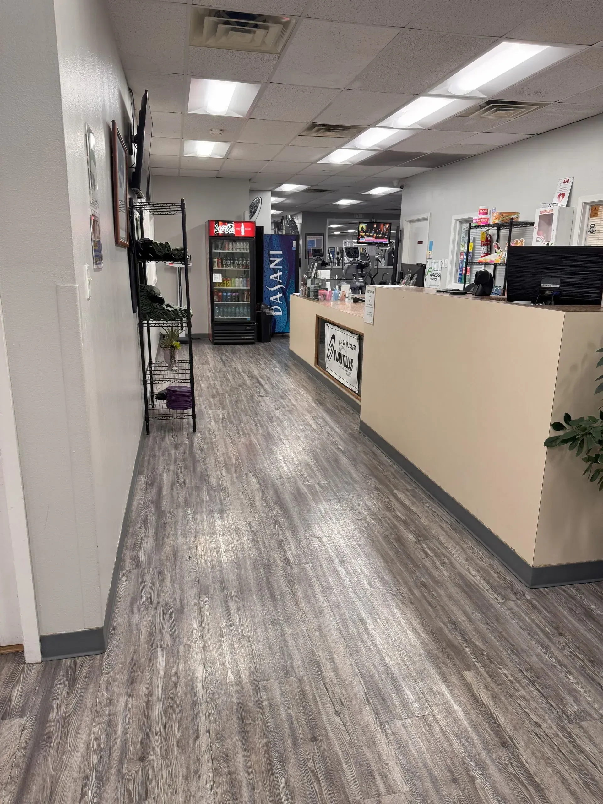 Interior view of a business hallway with a reception desk, cooler, and shelves. Gray wood-look floor.