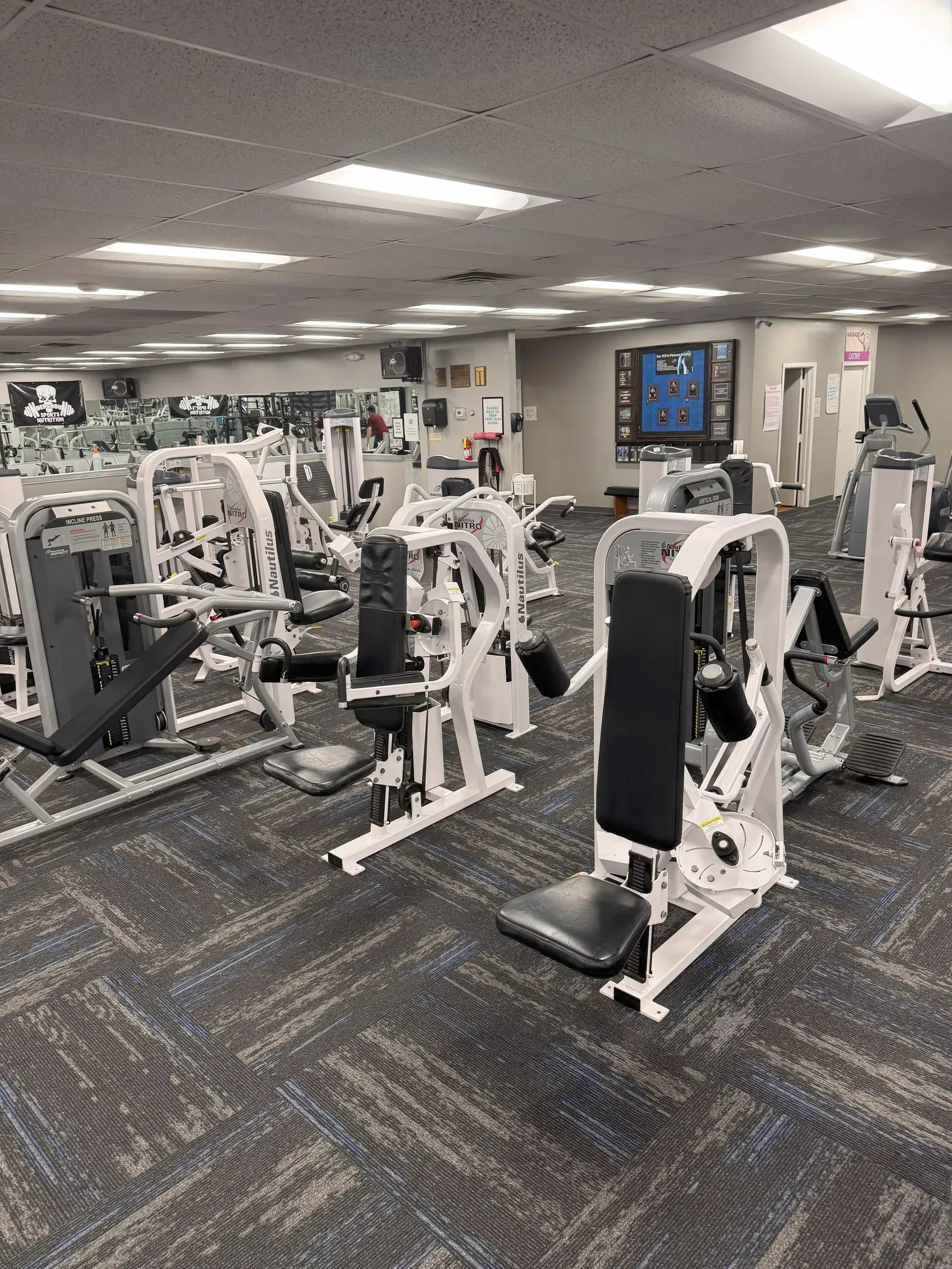 Gym interior with exercise machines on patterned carpet, under fluorescent lights.