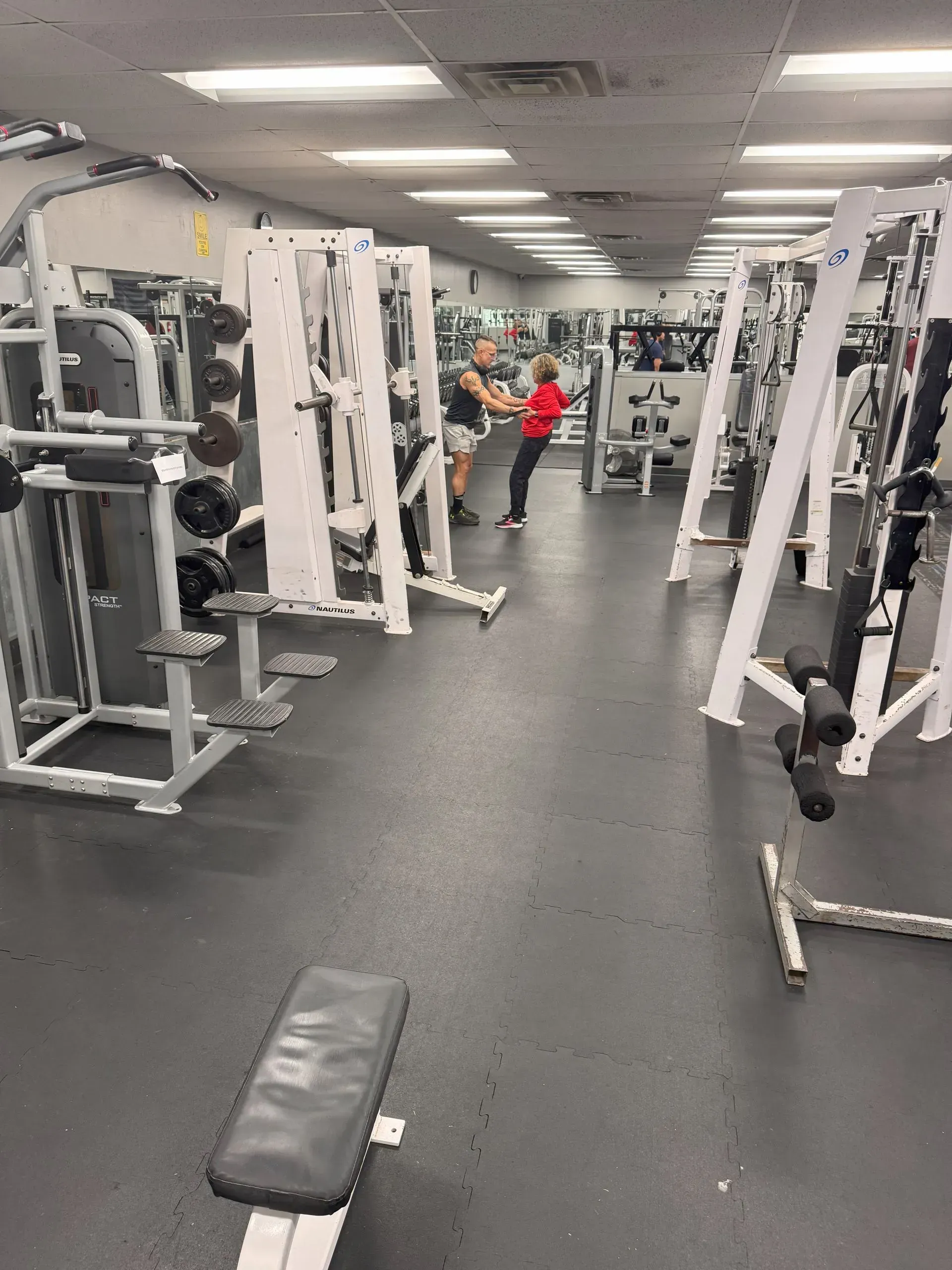 Gym interior with exercise machines. Two people use equipment; gray and white machines line the room.