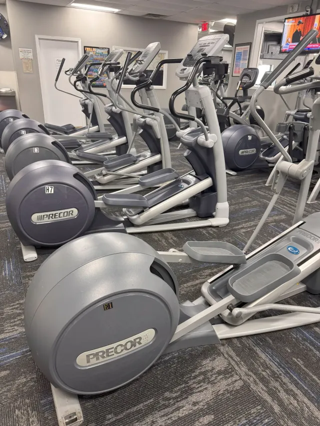 Row of Precor elliptical machines in a gym with a door and TVs visible in the background.