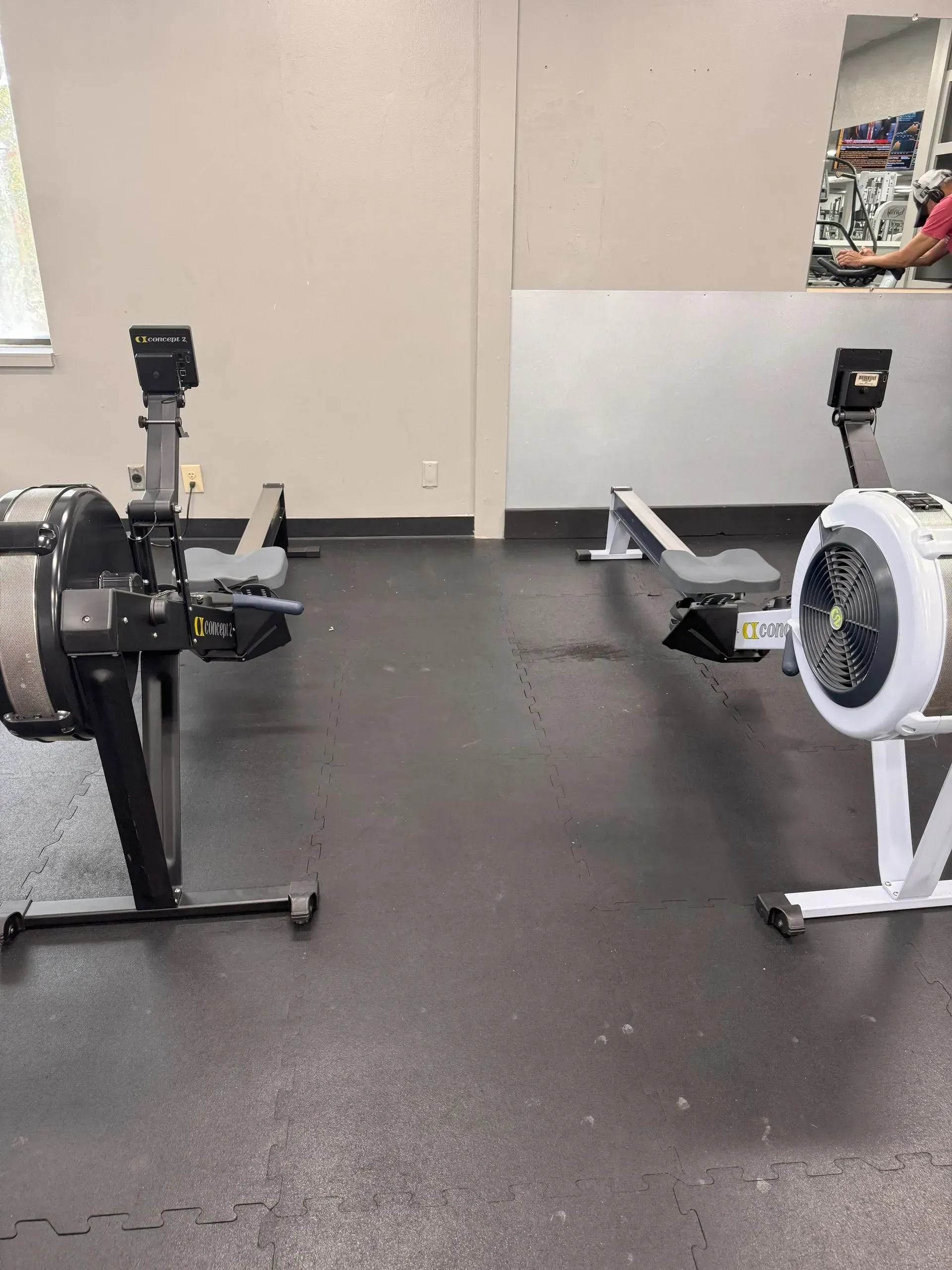 Two rowing machines on a black floor in a gym setting. One is black, the other white.