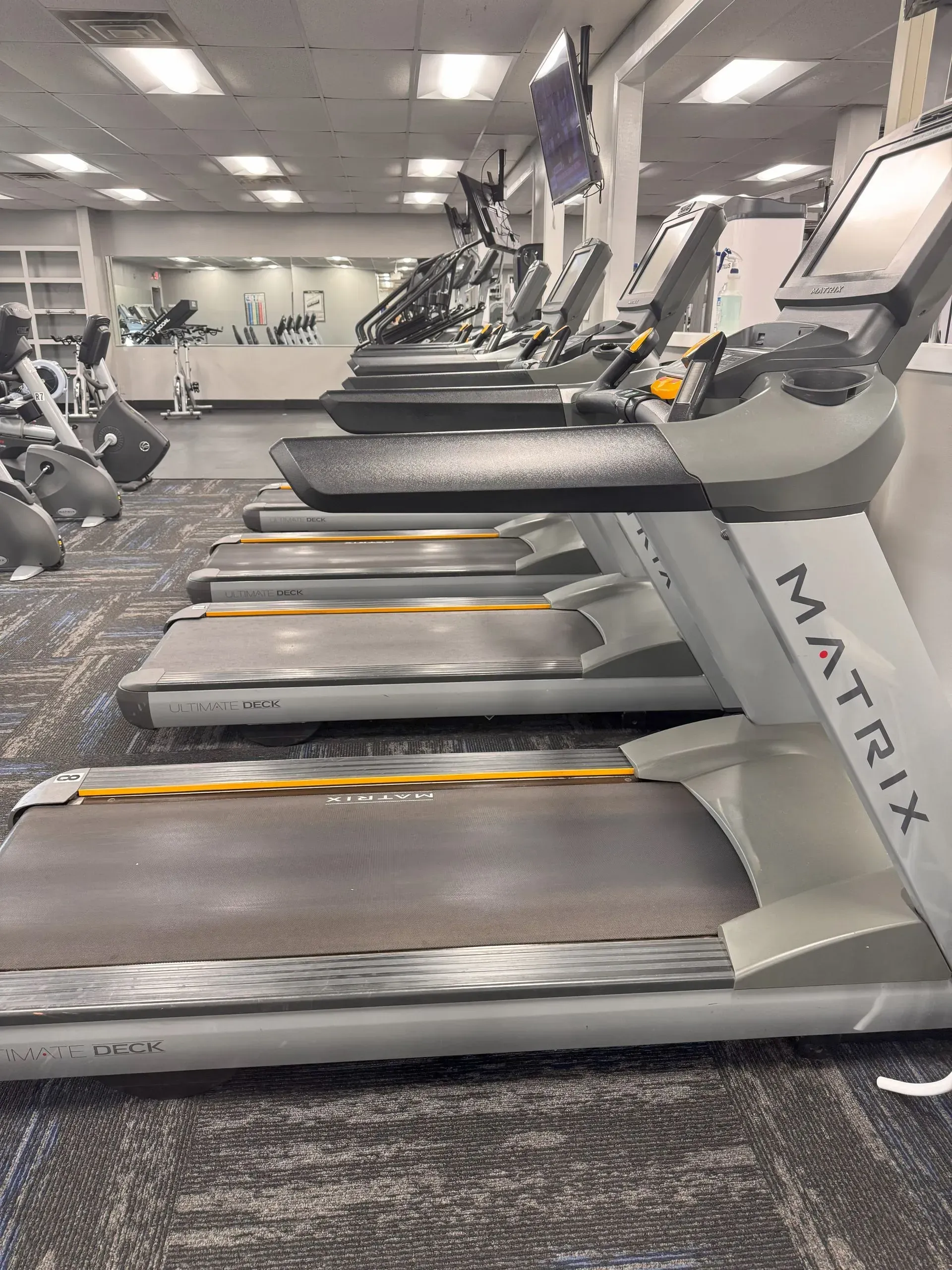 Row of treadmills in a gym setting with various exercise machines. Gray, yellow, and silver.