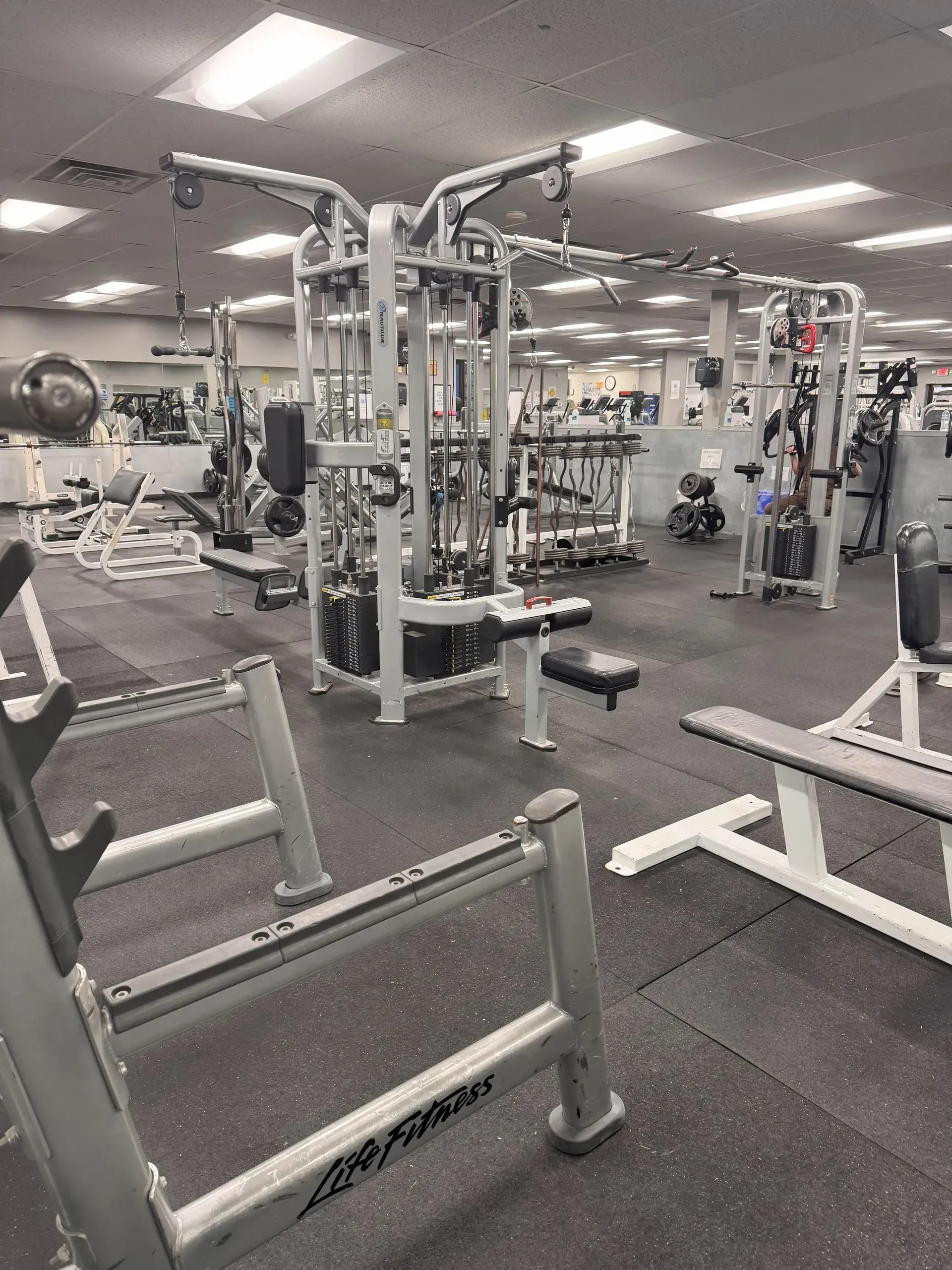Gym interior with weight machines, benches, and a rack, all on a dark floor.