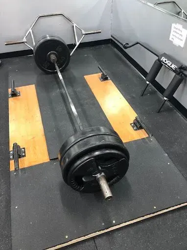 Barbell loaded with weight plates on a lifting platform, with a hex bar and Rogue supports in the background.