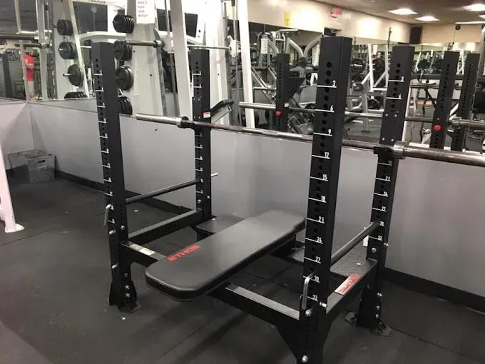 Weight bench with barbell on safety supports in a gym. Black equipment, gray walls.