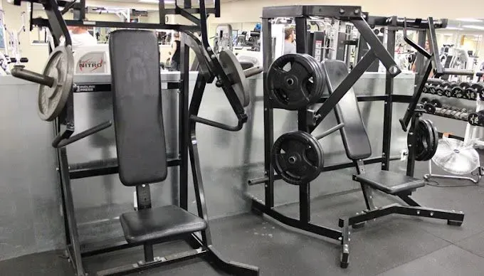 Two weight machines in a gym; a seated chest press and a shoulder press, both loaded with weights.